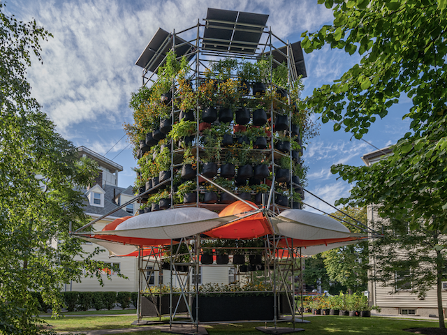 With solar panels at the top and white and orange inflatable pods at the bottom, a scaffolding structure nearly three stories high supports rings of potted plants 
